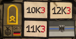 Nahaufnahme eines olivgrünen Militärrucksacks mit Abzeichen: dt. OR-7 Rang, 10K3, 11K3, 12K3 Codes u. ein dt. Thin Blue Line Patch.