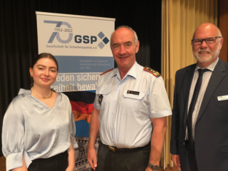 Eine junge Frau, ein Mann in Uniform und ein Mann im Anzug lächeln vor einem GSP Banner zum 70-jährigen Jubiläum.