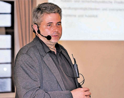 Ein Mann mit grauem Haar und Headset-Mikrofon spricht bei einer Präsentation. Er trägt einen dunklen Blazer und Hemd.