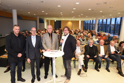 Fünf Männer stehen vor einem sitzenden Publikum älterer Menschen in einem Konferenzraum; einer hält ein Mikrofon.