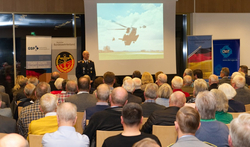Bundeswehroffizier präsentiert auf einer Leinwand fliegende Hubschrauber vor einem Publikum.