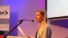 Eine blonde Frau spricht an einem Rednerpult mit Mikrofon und Papieren, vor einem lila Hintergrund und Banner.