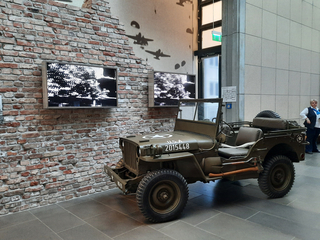 Ausgestellter Vintage-Militär-Jeep neben einer Ziegelwand mit Bildschirmen, Flugzeugsilhouetten und einer zuschauenden Frau.