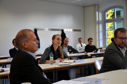 Junge Frau spricht oder hört aufmerksam in einem hellen Konferenzraum mit weiteren Teilnehmern an Tischen zu.