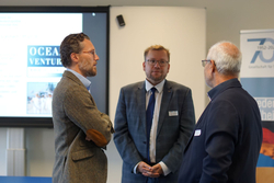 Drei Männer in Business-Kleidung diskutieren auf einer Veranstaltung. Im Hintergrund ein Bildschirm mit 'OCEAN VENTUR' und ein 70-Jahre-Jubiläumsbanner.