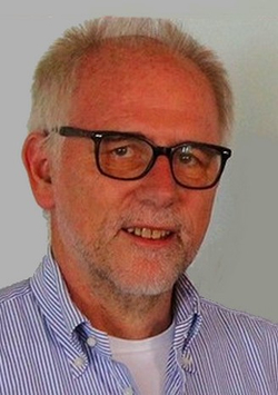 Lächelnder Mann mit grauen Haaren, schwarzer Brille und blau gestreiftem Hemd.