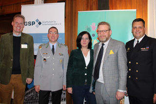 Fünf lächelnde Personen, darunter Militäroffiziere und Zivilisten, stehen vor GSP (Gesellschaft für Sicherheitspolitik) Bannern.