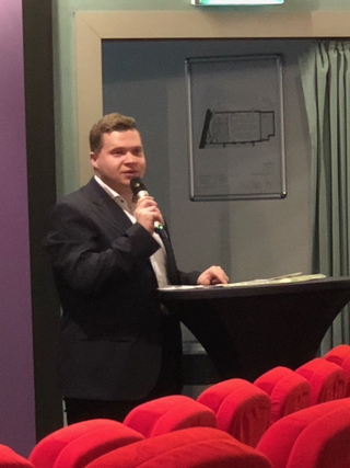 Ein Mann im dunklen Anzug spricht mit Mikrofon an einem Rednerpult in einem Auditorium mit roten Sitzen.