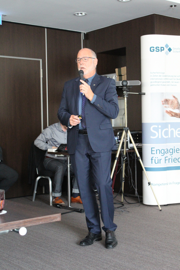 Mann im Anzug hält einen Vortrag und spricht in ein Mikrofon. Ein Banner mit "GSP" und "Sicherheit" ist im Hintergrund.
