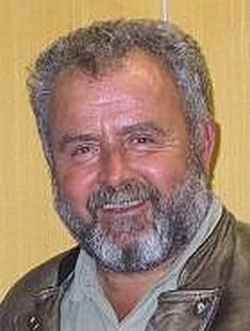 Porträt eines lächelnden Mannes mit grauem Bart und kurzen grauen Haaren, eine Jacke tragend.
