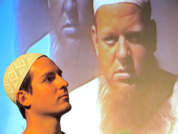 Junger Mann mit weißer Kufi blickt auf die Projektion eines älteren Mannes mit Bart und Kufi.