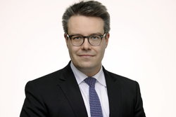 Porträt eines lächelnden Mannes mit Brille, dunklem Anzug und blau gemusterter Krawatte vor weißem Hintergrund.