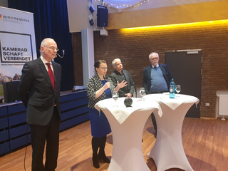 Eine Frau spricht am Mikrofon, umgeben von drei Männern und Stehtischen, bei einer Veranstaltung des Reservistenverbandes.