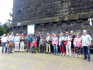 Eine Gruppe meist erwachsener Personen posiert vor einer massiven dunklen Steinmauer, wahrscheinlich einem historischen Denkmal oder Damm.