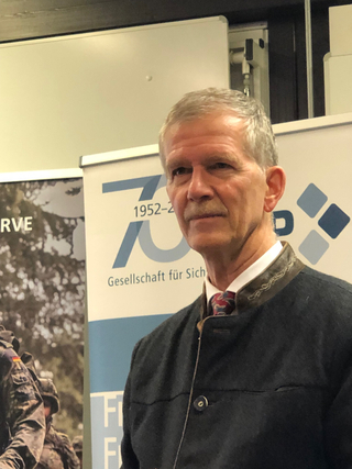 Älterer Mann mit Schnurrbart in Trachtenjacke bei Sicherheitspolitik-Event, 70-jähriges Jubiläumsbanner im Hintergrund.