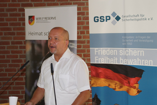 Sprechender Mann im weißen Hemd vor Bannern des Reservistenverbandes und der Gesellschaft für Sicherheitspolitik.