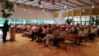 Ein Mann spricht an einem Pult vor einem sitzenden Publikum in einer großen Halle.