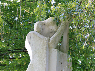 Verwitterte Steinskulptur einer nachdenklichen Figur mit gesenktem Kopf, umrahmt von grünen Trauerweidenblättern.