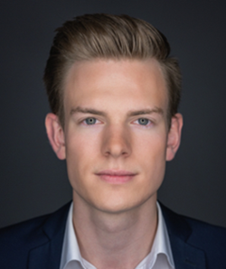 Porträt eines jungen Mannes mit blonden Haaren und blauen Augen, der einen dunklen Anzug und ein weißes Hemd trägt.