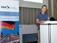 Ein Mann spricht an einem Pult neben einem Banner der Gesellschaft für Sicherheitspolitik (GSP) mit deutscher Flagge.