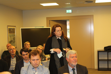 Eine Frau mit Brille und Maske in der Hand steht in einem Konferenzraum vor sitzenden Zuhörern.