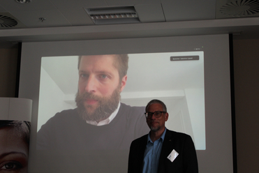 Ein Mann präsentiert in einem Raum vor einer Leinwand, die einen bärtigen Mann im Videoanruf/Präsentation zeigt.