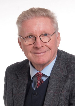 Porträt eines lächelnden älteren Mannes mit grauem Haar und Brille, in grauer Tweed-Jacke, dunkler Weste und roter Krawatte.
