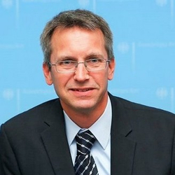 Porträt eines Mannes mittleren Alters mit Brille, dunklem Anzug und gestreifter Krawatte vor hellblauem Hintergrund.