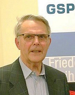 Porträt eines älteren Mannes mit Brille, hellem Haar und dunklem Sakko, leicht lächelnd vor einem GSP- und Fried-Hintergrund.