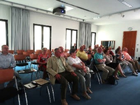 Eine Gruppe von Erwachsenen, hauptsächlich ältere Menschen, sitzt in Reihen in einem hellen Konferenzraum mit Beamer.