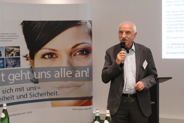 Ein Mann in einem Anzug spricht in ein Mikrofon bei einer Präsentation, im Hintergrund ein deutsches Banner mit Frauengesicht.