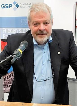 Ein bärtiger Mann mit Brille steht an einem Rednerpult mit Mikrofon vor einem Banner der 'Gesellschaft für Sicherheitspolitik'.
