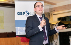 Ein Mann in Anzug und Brille spricht in ein Mikrofon, gestikulierend, vor einem GSP-Banner und der deutschen Flagge.