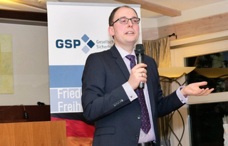 Ein Mann in Anzug und Brille spricht in ein Mikrofon, gestikulierend, vor einem GSP-Banner und der deutschen Flagge.