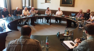 Eine Gruppe von Personen, darunter einige in Uniform, sitzt an einem großen U-förmigen Tisch in einem Besprechungsraum.