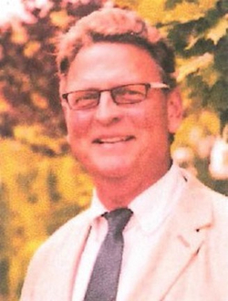 Nahaufnahme eines lächelnden Mannes mit Brille, heller Jacke und dunkler Krawatte, vor unscharfem Hintergrund im Freien.