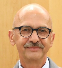 Porträt eines lächelnden, glatzköpfigen Mannes mit Brille und Schnurrbart.