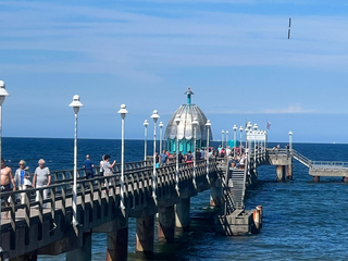 Blick auf die Seebrücke Zinnowitz mit ihrer charakteristischen Tauchgondel und vielen Spaziergängern bei Sonnenschein.