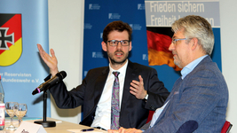 Zwei Männer diskutieren auf einem Podium. Einer gestikuliert sprechend, der andere hört zu. Banner mit Bundeswehr, Frieden, Freiheit.
