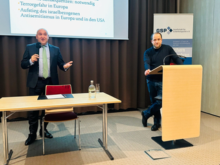 Mann im Anzug präsentiert mit Mikrofon auf GSP-Sicherheitspolitik-Konferenz, anderer Mann steht am Pult.