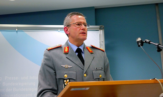 Deutscher General Bodemann in Uniform spricht an einem Rednerpult auf einer Pressekonferenz, mit einem Regierungsbanner dahinter.