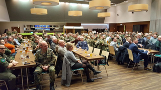 Eine große Gruppe von Menschen, viele in Militäruniformen, sitzen an Tischen in einem Saal und verfolgen eine Veranstaltung.