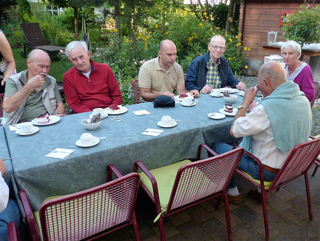Sechs Personen, überwiegend ältere Erwachsene, genießen Kaffee und Kuchen an einem Tisch im Garten.