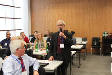 Ein Mann spricht auf einer Konferenz ins Mikrofon, während Zuhörer in einem hellen Raum mit Holzvertäfelung lauschen.