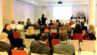 Zuhörer in einem Konferenzraum lauschen einem Vortragenden und einem Podium. Eine Präsentation ist auf einer Leinwand zu sehen.