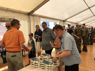 Menschen verschiedener Altersgruppen, darunter auch Soldaten, an einem Stand unter einem Zelt, wo Bücher zum Thema Sicherheitspolitik ausgelegt sind.
