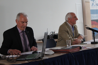 Zwei Männer an einem Konferenztisch, einer auf einen Laptop konzentriert, der andere, ein älterer Mann, lächelt mit Mikrofon.
