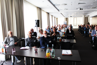 Eine Gruppe von Teilnehmern sitzt aufmerksam an Tischen in einem Konferenzraum mit Getränken und Notizbüchern.