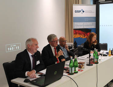 Vier Teilnehmer einer GSP-Sicherheitspolitik-Konferenz, ein Mann spricht ins Mikrofon, im Hintergrund ein Banner.