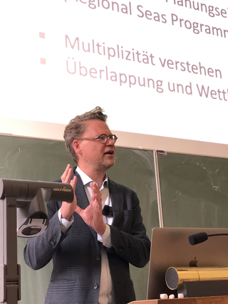 Mann mit Brille hält Vortrag, gestikulierend vor einer Präsentation über Multiplizität und Überlappung.